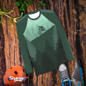 May include: A long-sleeved shirt with a green and white forest design. The shirt features a silhouette of a person riding a mountain bike through the trees.