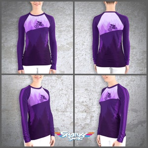 Peut inclure: Chemise &agrave; manches longues violette avec un motif repr&eacute;sentant une personne faisant du v&eacute;lo de montagne dans une for&ecirc;t. La chemise a une bande blanche sur les manches.