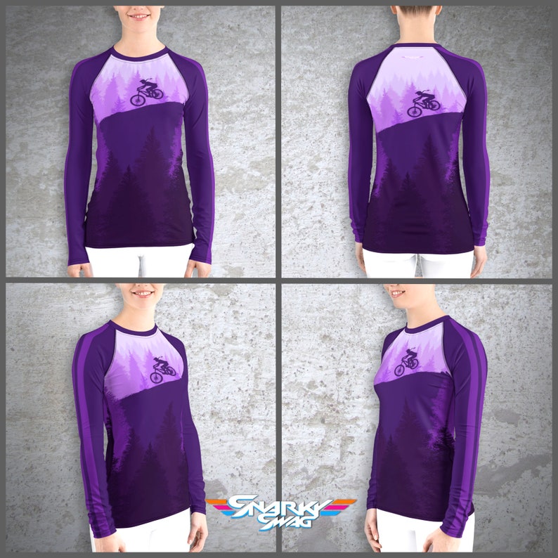 Peut inclure: Chemise &agrave; manches longues violette avec un motif repr&eacute;sentant une personne faisant du v&eacute;lo de montagne dans une for&ecirc;t. La chemise a une bande blanche sur les manches.
