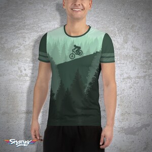 May include: A green t-shirt featuring a mountain biking scene. The design includes a silhouette of a cyclist on a bike, set against a backdrop of pine trees and a gradient sky. The shirt has a crew neck and striped sleeves.