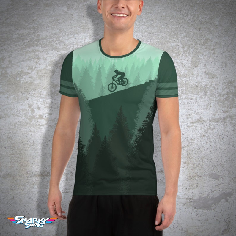 May include: A green t-shirt featuring a mountain biking scene. The design includes a silhouette of a cyclist on a bike, set against a backdrop of pine trees and a gradient sky. The shirt has a crew neck and striped sleeves.