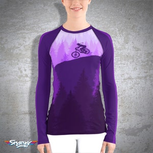 Peut inclure: T-shirt &agrave; manches longues violette avec une silhouette d'une personne faisant du v&eacute;lo de montagne dans une for&ecirc;t. Le t-shirt a un panneau blanc sur le devant et des manches violettes.