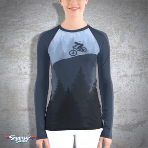 Peut inclure: Un t-shirt &agrave; manches longues avec un motif bleu fonc&eacute; et bleu clair. Le t-shirt pr&eacute;sente une silhouette d'une personne faisant du v&eacute;lo de montagne dans une for&ecirc;t.