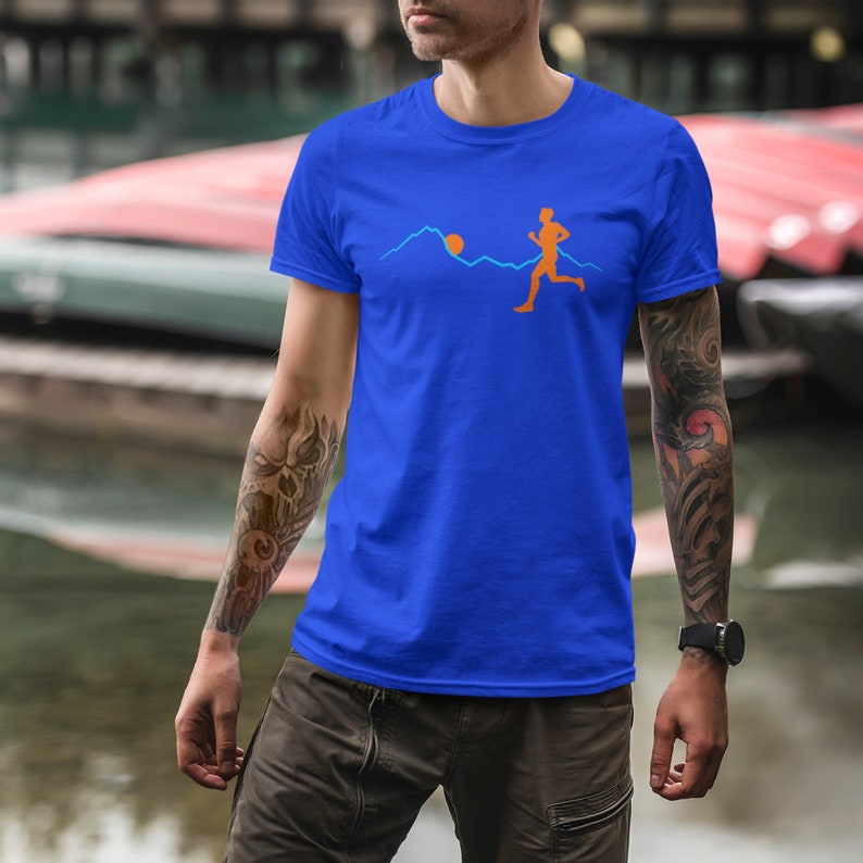May include: A royal blue t-shirt featuring a graphic of a person running in front of a mountain range with a sun rising behind the mountains. The graphic is in orange and blue.