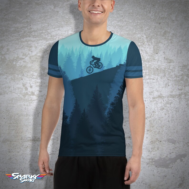May include: A blue t-shirt featuring a mountain biking scene. The design shows a cyclist on a bike against a backdrop of a forest and sky in shades of blue. The shirt has a crew neck and striped sleeves.