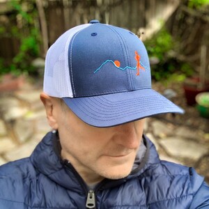 Puede incluir: Una gorra de camionero azul y blanca con un dise&ntilde;o bordado de una persona corriendo frente a una cordillera con el sol poni&eacute;ndose detr&aacute;s.