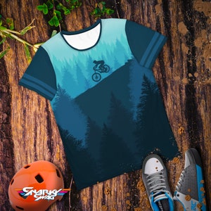 May include: A blue t-shirt with a mountain biking graphic. The shirt features a forest scene in shades of blue and a cyclist silhouette. The sleeves have blue stripes. An orange helmet and athletic shoes are also in the image.