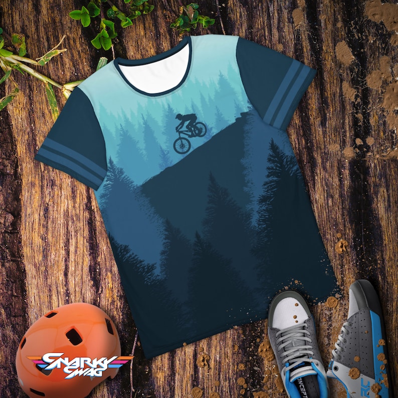 May include: A blue t-shirt with a mountain biking graphic. The shirt features a forest scene in shades of blue and a cyclist silhouette. The sleeves have blue stripes. An orange helmet and athletic shoes are also in the image.