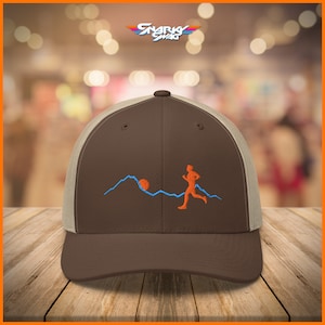 Puede incluir: Gorra de b&eacute;isbol marr&oacute;n y beige con una parte trasera de malla marr&oacute;n. La parte delantera de la gorra presenta un dise&ntilde;o bordado de una persona corriendo por una cordillera con un sol en el fondo.