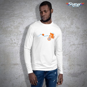 May include: White long-sleeve t-shirt with a graphic of a person riding a bicycle up a mountain with a sun in the background. The text "SNARK SWAG" is in the top right corner of the image.