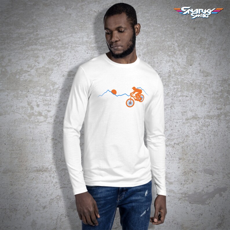 May include: White long-sleeve t-shirt with a graphic of a person riding a bicycle up a mountain with a sun in the background. The text "SNARK SWAG" is in the top right corner of the image.