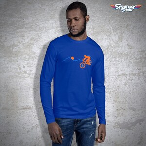 May include: A royal blue long-sleeve t-shirt with an orange silhouette of a person riding a mountain bike over a mountain range with a setting sun. The text "SNARKY SWAG" is in the upper right corner of the image.