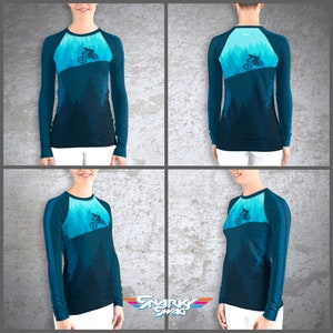 Peut inclure: Un t-shirt &agrave; manches longues avec un d&eacute;grad&eacute; de couleurs bleu et turquoise. Le t-shirt pr&eacute;sente une silhouette d'une personne faisant du v&eacute;lo de montagne dans une for&ecirc;t. Le t-shirt a une conception de manche raglan avec une bande bleu fonc&eacute; sur l'&eacute;paule.