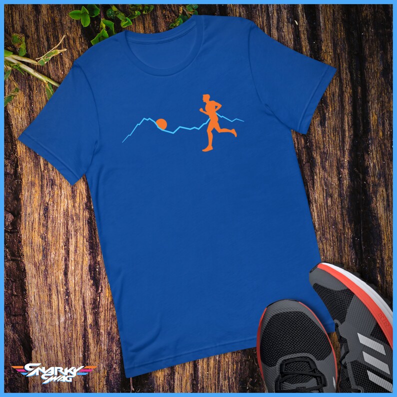 May include: A royal blue t-shirt featuring a graphic of a person running across a mountain range with a sun rising in the background.