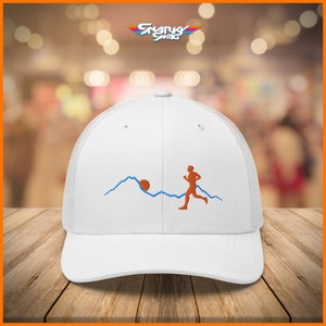 Puede incluir: Gorra de camionero blanca con un dise&ntilde;o bordado en azul y naranja de una persona corriendo en una monta&ntilde;a con una pelota. El dise&ntilde;o est&aacute; en la parte delantera de la gorra.