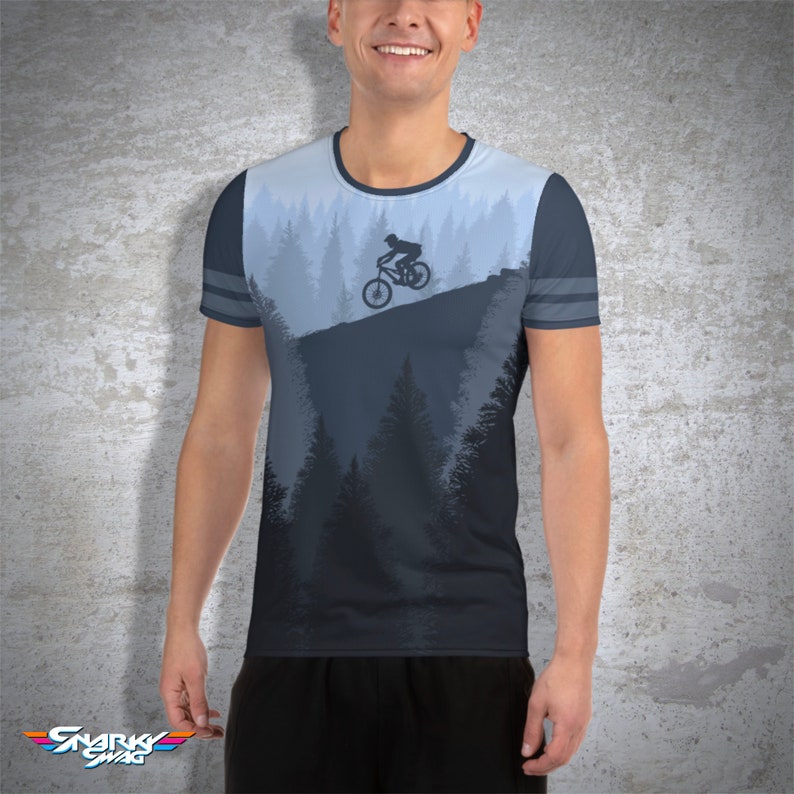 May include: A blue and gray t-shirt featuring a mountain biking design. The shirt has a crew neck and short sleeves with gray stripes. The graphic depicts a cyclist on a mountain trail with a forest backdrop. The Snarky Swag logo is at the bottom.