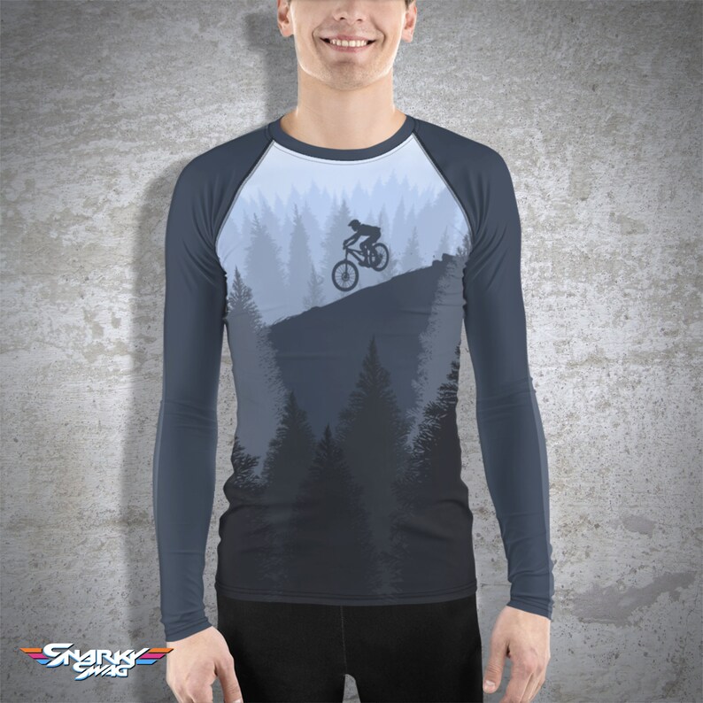 May include: A long-sleeved shirt with a dark blue base color and a light blue raglan sleeve design. The shirt features a graphic of a mountain biker riding through a forest.