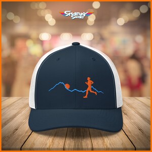 Puede incluir: Una gorra de camionero azul marino y blanca con una figura corriendo y una silueta de monta&ntilde;a bordada en la parte delantera. El bordado es de color naranja y azul.