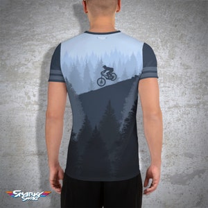 May include: A light blue and dark grey t-shirt featuring a mountain biking scene. The design shows a cyclist on a mountain slope with a forest backdrop. The sleeves have grey stripes. The brand logo "Snarky Swag" is at the bottom.