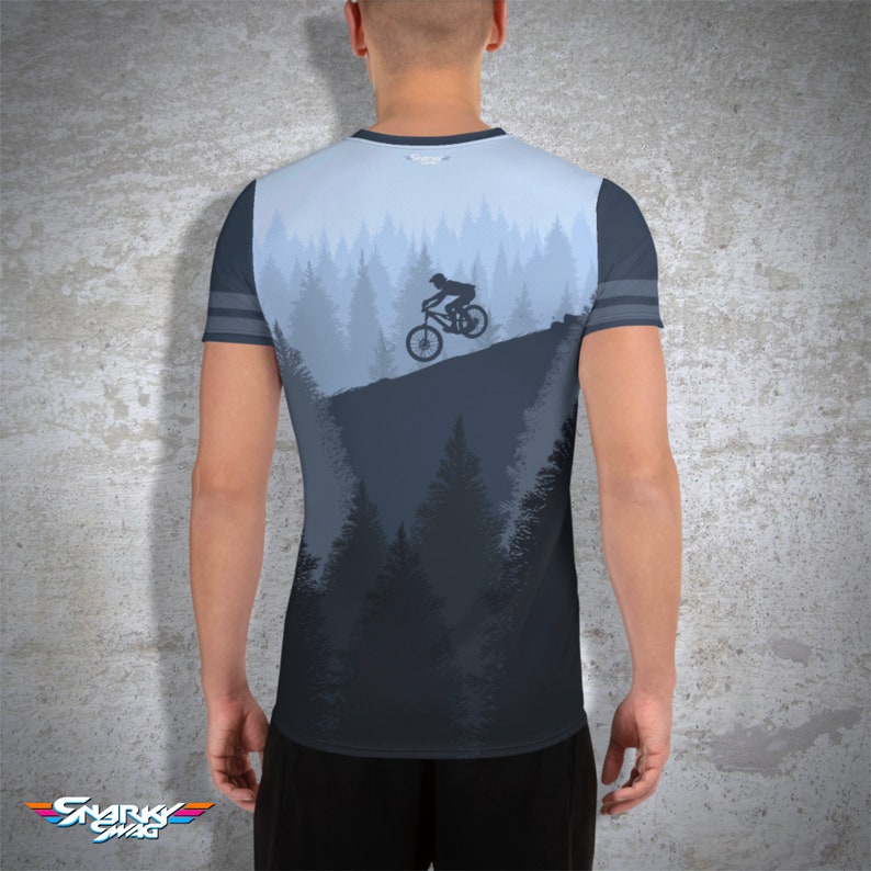 May include: A light blue and dark grey t-shirt featuring a mountain biking scene. The design shows a cyclist on a mountain slope with a forest backdrop. The sleeves have grey stripes. The brand logo "Snarky Swag" is at the bottom.