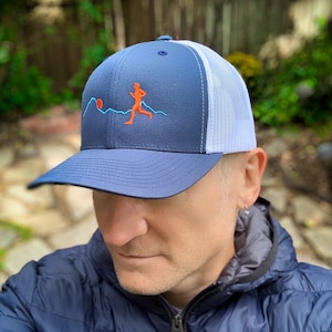 Puede incluir: Una gorra de camionero azul marino y blanca con una malla blanca en la parte posterior. La gorra presenta un dise&ntilde;o bordado de una persona corriendo frente a una cordillera con un sol rojo.
