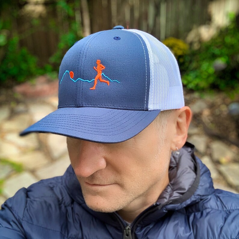 Puede incluir: Una gorra de camionero azul marino y blanca con una malla blanca en la parte posterior. La gorra presenta un dise&ntilde;o bordado de una persona corriendo frente a una cordillera con un sol rojo.