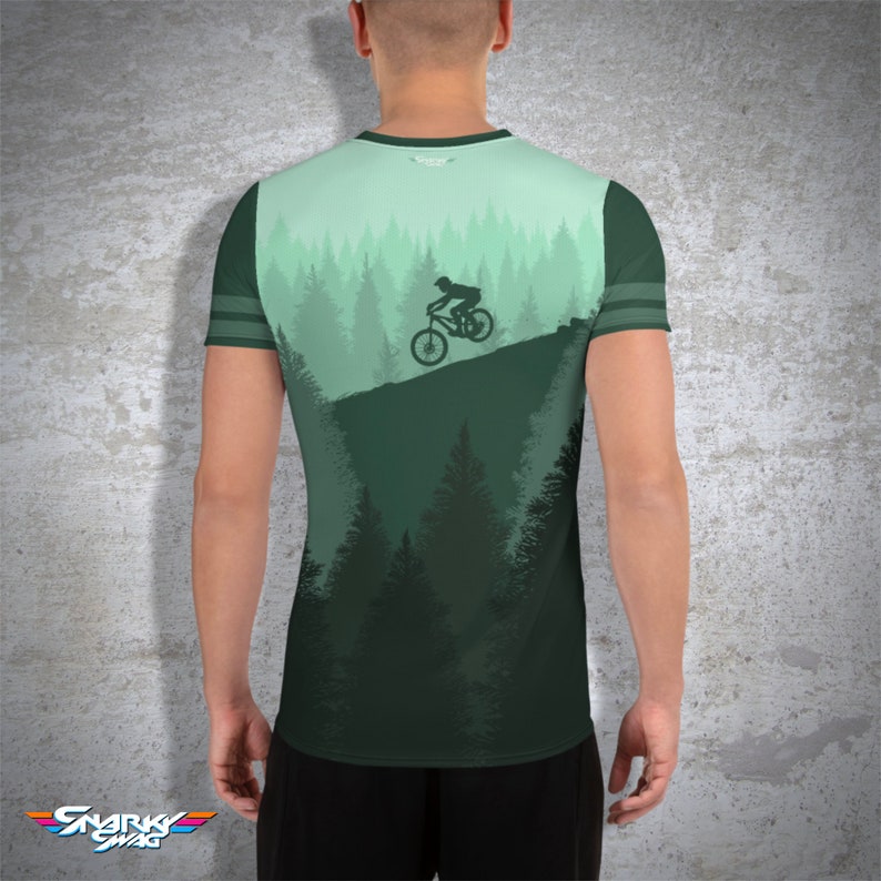 May include: A green and teal athletic t-shirt featuring a mountain biking design. The shirt has a silhouette of a cyclist on a mountain slope, with a forest backdrop. The sleeves have dark green stripes. The Snarky Swag logo is at the bottom.