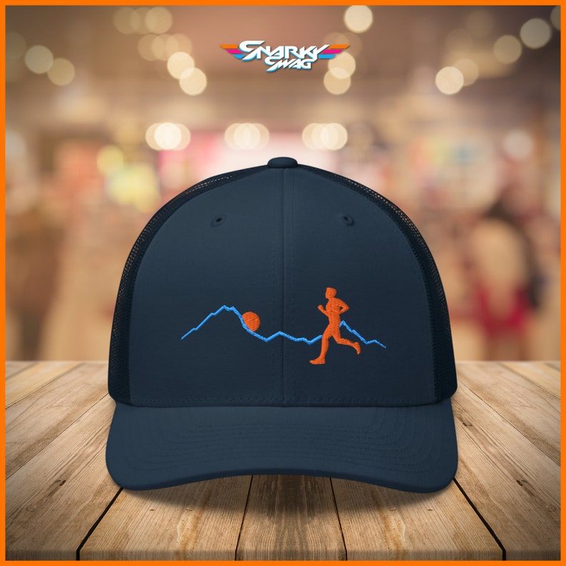 Puede incluir: Una gorra de camionero azul marino con una parte trasera de malla negra. La gorra presenta un dise&ntilde;o bordado de una persona corriendo frente a una cordillera con el sol poni&eacute;ndose detr&aacute;s de las monta&ntilde;as.