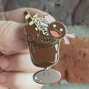 Puede incluir: Un pin de esmalte de un sundae de chocolate con un contorno dorado. El pin presenta una copa de sundae marrón llena de helado de chocolate, crema batida, chispas y un hombre de jengibre sonriente con acentos rosados. Un accesorio decorativo.