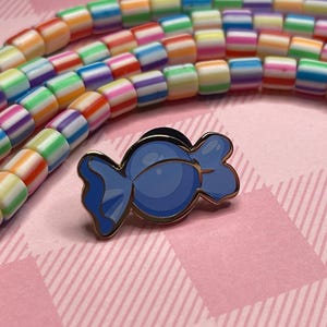Può includere: Spilla in smalto a forma di caramella blu con contorno dorato. La spilla è posta su uno sfondo a scacchi rosa e bianco, con perline a righe colorate sullo sfondo. Un accessorio divertente.