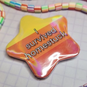 Puede incluir: Una insignia en forma de estrella con el texto "I survived homestuck". La insignia tiene un degradado de amarillo a naranja y un brillo holográfico. Collares de cuentas coloridas en el fondo.