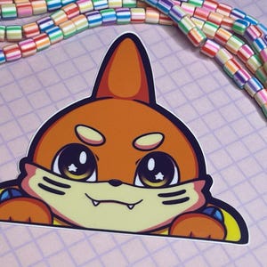 May include: A die-cut sticker featuring a cartoon fox with orange fur, large eyes with star accents, and a smiling expression. The sticker has yellow and blue accents. Colorful, striped beads are in the background.