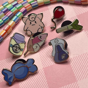 May include: A collection of enamel pins featuring various designs, including a pink Clefairy, a red balloon, a green leek, a blue candy, and other character-themed pins. The pins have gold-colored borders and are set against a pink plaid background.