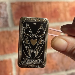 Könnte beinhalten: Ein goldfarbener rechteckiger Ansteckpin mit dem Design der Tarotkarte Die Liebenden. Der Pin hat einen schwarzen Hintergrund mit zwei drachenartigen Figuren, die sich gegenüberstehen, einem Herz in der Mitte und den Worten "THE LOVERS" unten.