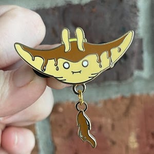 May include: A yellow and brown enamel pin featuring a cartoon manta ray design with a smiling face and dripping brown "sauce". A small, dangling charm hangs below. The pin is held against a brick background.