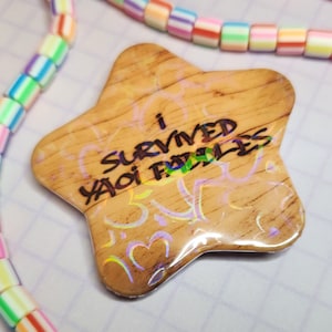 May include: A star-shaped wooden pin with the text "I SURVIVED YAOI FANFICS" in black. The pin has a holographic effect and is surrounded by a colorful beaded necklace.