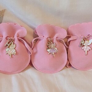 May include: Three light pink velvet drawstring pouches, each featuring a metal charm attached to the cord. The charms include a silver-toned letter H, S, or B paired with a small, colourful enamel fairy figure in shades of pink, purple, and white.
