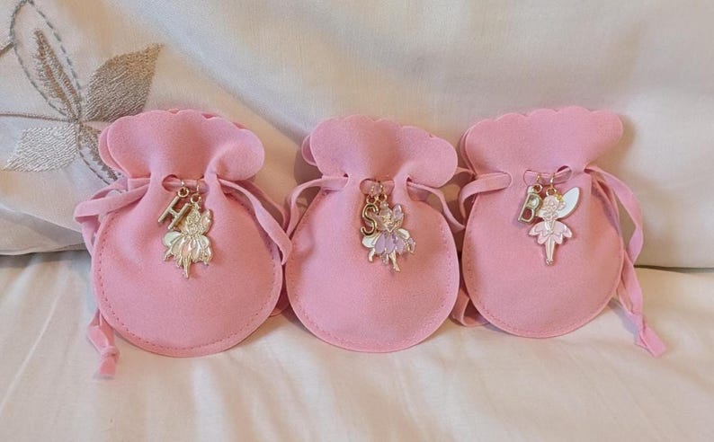 May include: Three light pink velvet drawstring pouches, each featuring a metal charm attached to the cord. The charms include a silver-toned letter H, S, or B paired with a small, colourful enamel fairy figure in shades of pink, purple, and white.