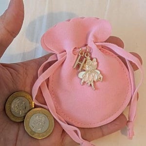 May include: A small, light pink felt drawstring pouch featuring a gold-toned metal charm of the letter H and a fairy figure. Two silver and gold-toned coins are positioned next to the pouch, providing a sense of scale for the small accessory.
