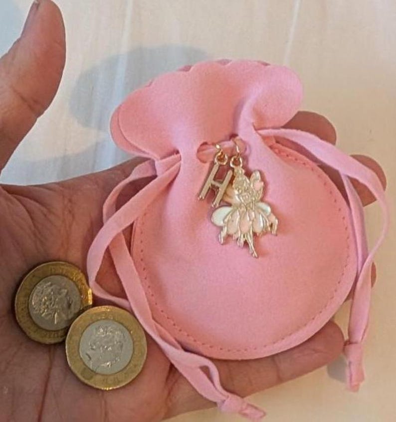May include: A small, light pink felt drawstring pouch featuring a gold-toned metal charm of the letter H and a fairy figure. Two silver and gold-toned coins are positioned next to the pouch, providing a sense of scale for the small accessory.