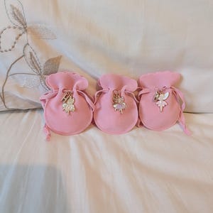 May include: Three small, round, light pink drawstring pouches made of a soft fabric. Each pouch features a metal charm attached to the drawstring, depicting a fairy figure paired with a single letter charm: H, S, and B respectively.