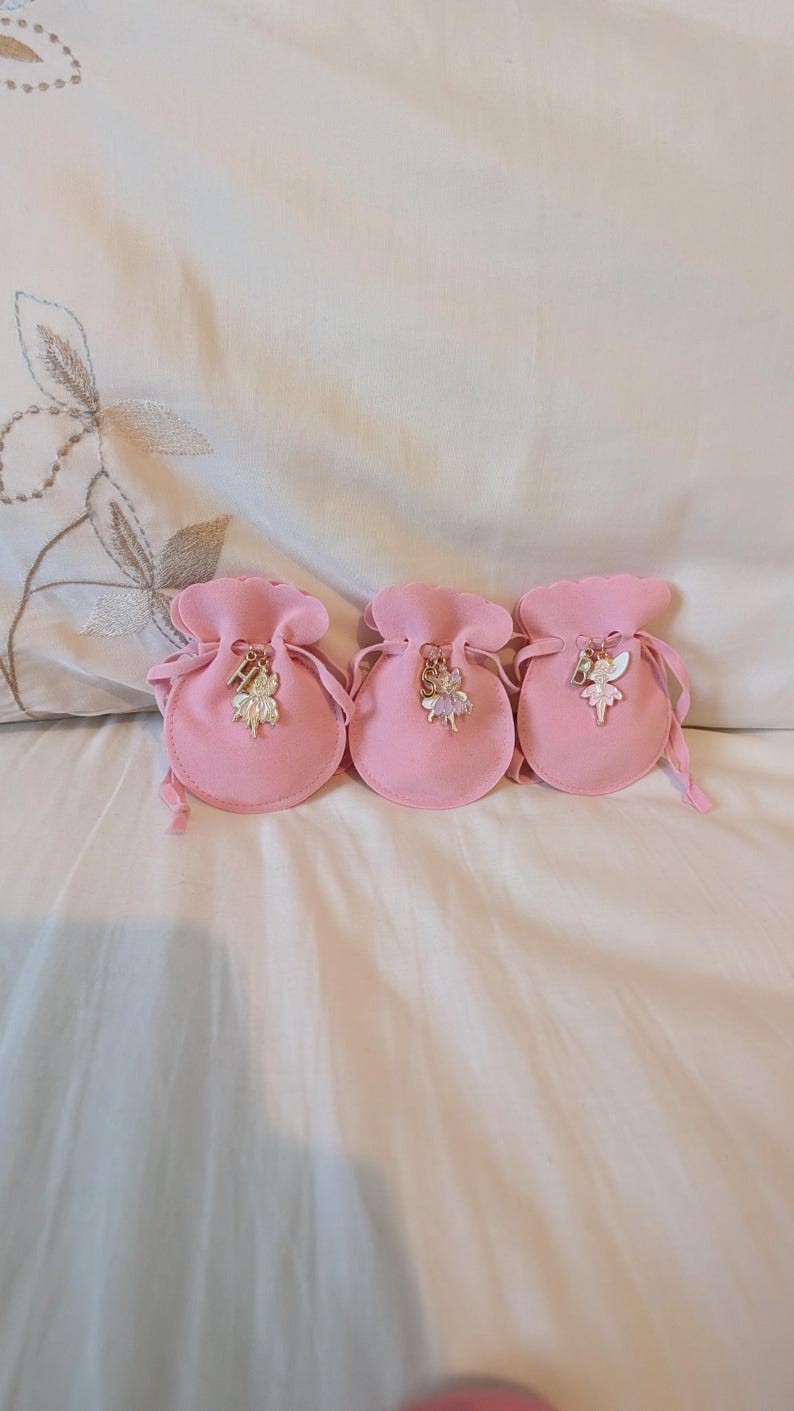 May include: Three small, round, light pink drawstring pouches made of a soft fabric. Each pouch features a metal charm attached to the drawstring, depicting a fairy figure paired with a single letter charm: H, S, and B respectively.
