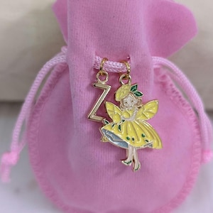 May include: A pink velvet drawstring pouch featuring two gold-toned metal charms attached to the cord. One charm is the letter Z, and the other is a figure wearing a yellow dress and hat with green accents, suspended from a gold jump ring.