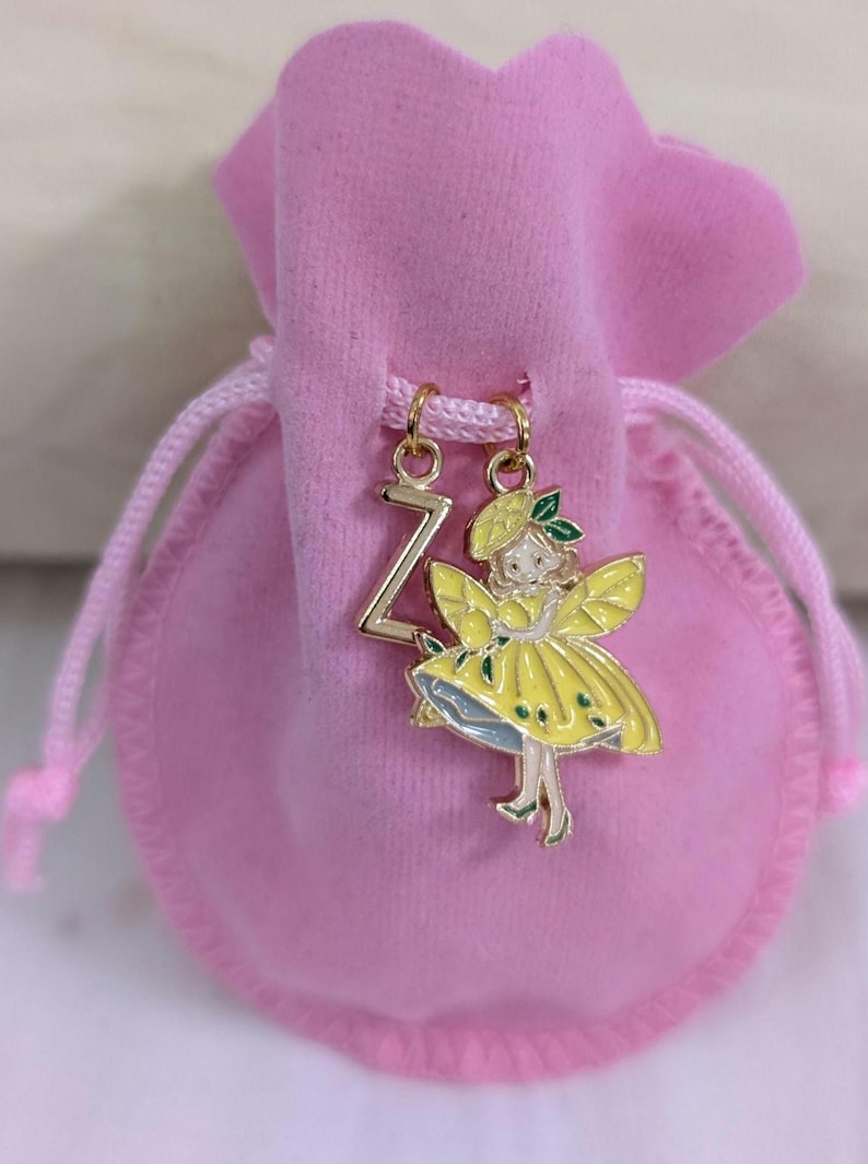 May include: A pink velvet drawstring pouch featuring two gold-toned metal charms attached to the cord. One charm is the letter Z, and the other is a figure wearing a yellow dress and hat with green accents, suspended from a gold jump ring.