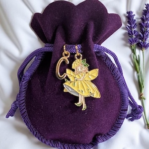 May include: Gold-coloured fairy earrings with yellow enamel wings and dress, and a gold letter "C". The earrings are presented with a purple velvet pouch and lavender sprigs.