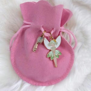 May include: A soft pink drawstring pouch featuring two metal charms attached to the cord. One charm is a small hairbrush, and the other is a fairy figure with white wings and a light green dress, both finished with gold-toned metal edges.