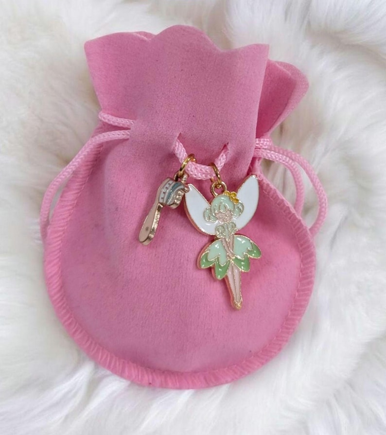 May include: A soft pink drawstring pouch featuring two metal charms attached to the cord. One charm is a small hairbrush, and the other is a fairy figure with white wings and a light green dress, both finished with gold-toned metal edges.
