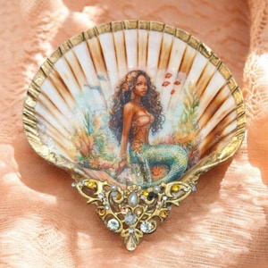 May include: A decorative scallop shell featuring a painted illustration of a mermaid with long dark hair and a green tail, surrounded by underwater flora. The shell edge is painted gold, and the base is adorned with gold metal filigree and crystals.