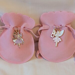 May include: Two light pink drawstring pouches made of a soft fabric. Each pouch features a gold-toned metal charm attached to the drawstring. One charm is a letter S with a purple fairy, and the other is a letter B with a white and pink fairy.