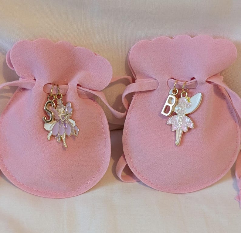 May include: Two light pink drawstring pouches made of a soft fabric. Each pouch features a gold-toned metal charm attached to the drawstring. One charm is a letter S with a purple fairy, and the other is a letter B with a white and pink fairy.